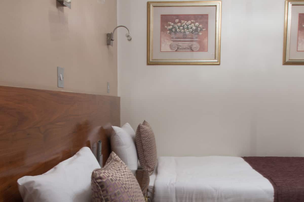 A hotel room with two beds, each with white linens and patterned pillows. There is a wooden headboard on the left bed, and a wall-mounted light fixture above the beds.