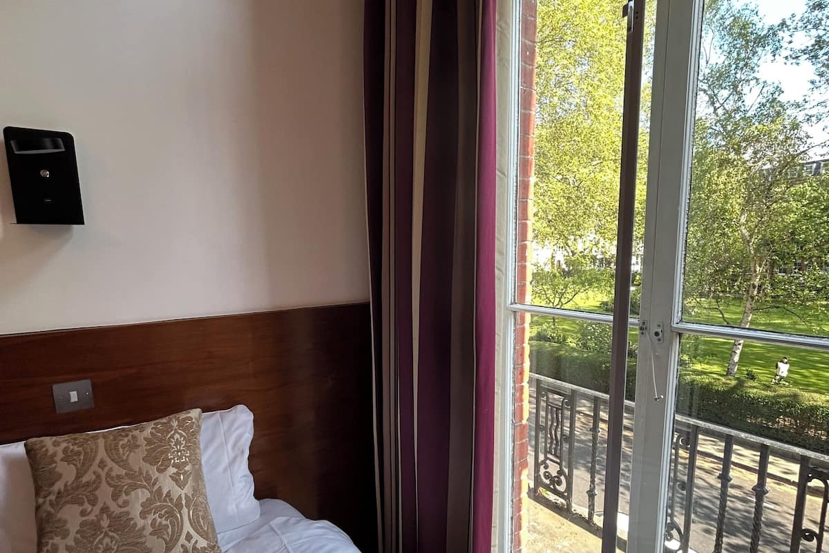 A hotel room with a bed, a wall-mounted control panel, and a view of a balcony with a railing and greenery.