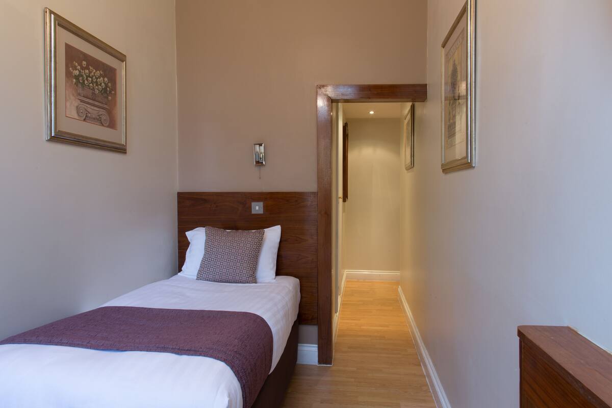 A hotel room with a bed, a wooden headboard, a nightstand, a framed picture on the wall, and a hallway leading to another room.