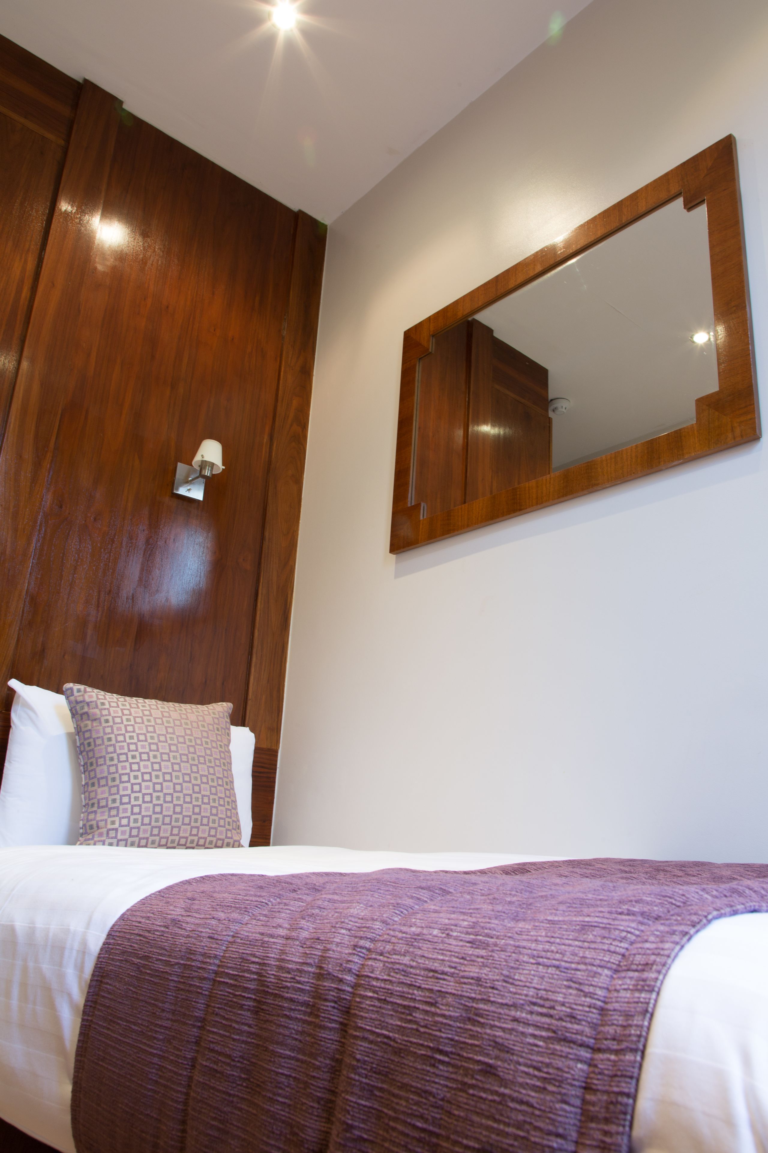 Une chambre d’hôtel avec une armoire en bois, un grand miroir, un lit avec une literie blanche et un plaid violet.