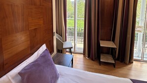 A hotel room with a wooden headboard, a bed with white and purple bedding, a small table, a chair, and a balcony with curtains. - Garden View Hotel (London)