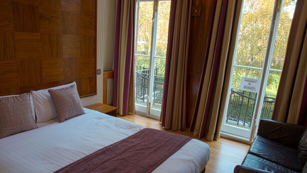 A hotel room with a large bed, a wooden headboard, a balcony with a view of trees, and a sign warning about the balcony. - Garden View Hotel (London)