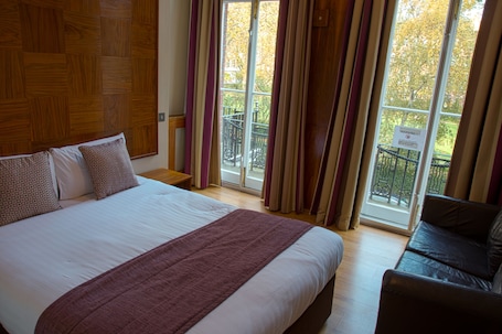 A hotel room with a large bed, a wooden headboard, a balcony with a view of trees, and a sign warning about the balcony.