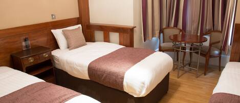 A hotel room with two beds, a wooden headboard, a bedside table with a lamp, a small table with chairs, and a framed picture on the wall.