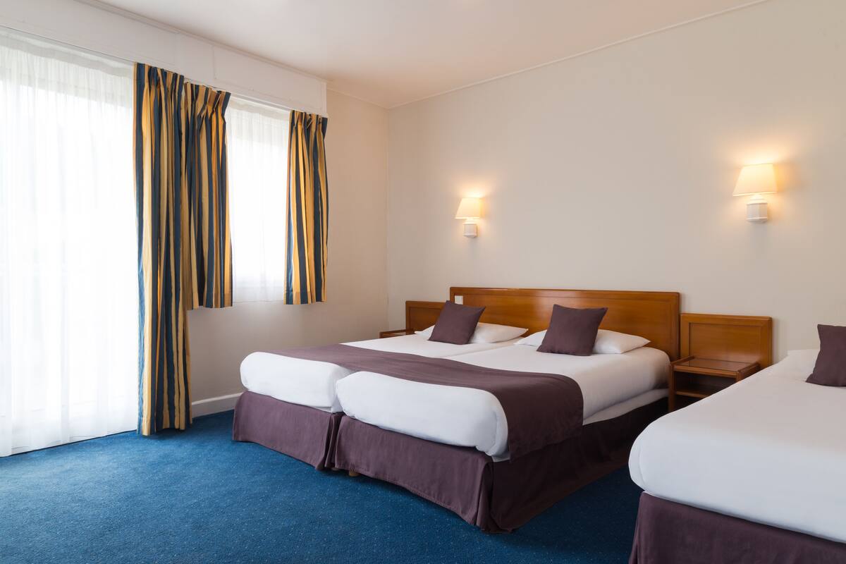A hotel room with two beds, a large window with curtains, and two wall-mounted lamps.