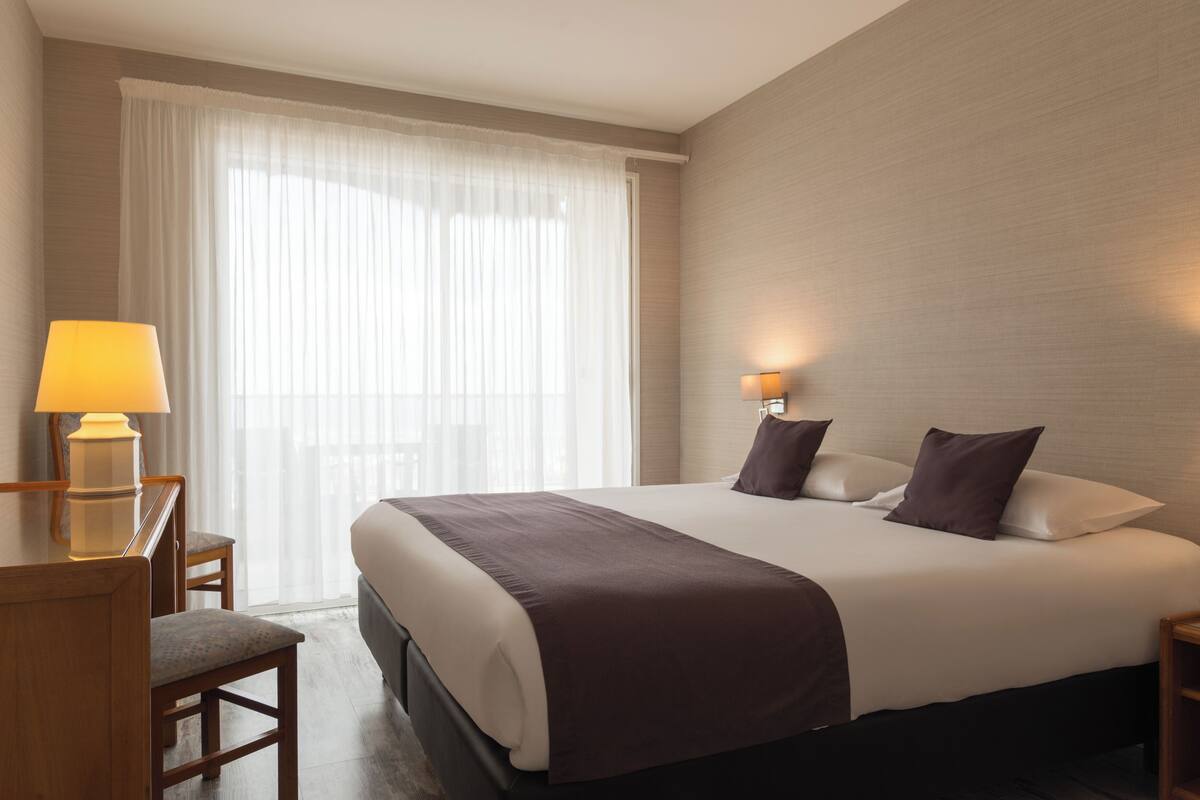 A hotel room with a large bed, two bedside lamps, a desk with a chair, and a window with sheer curtains.