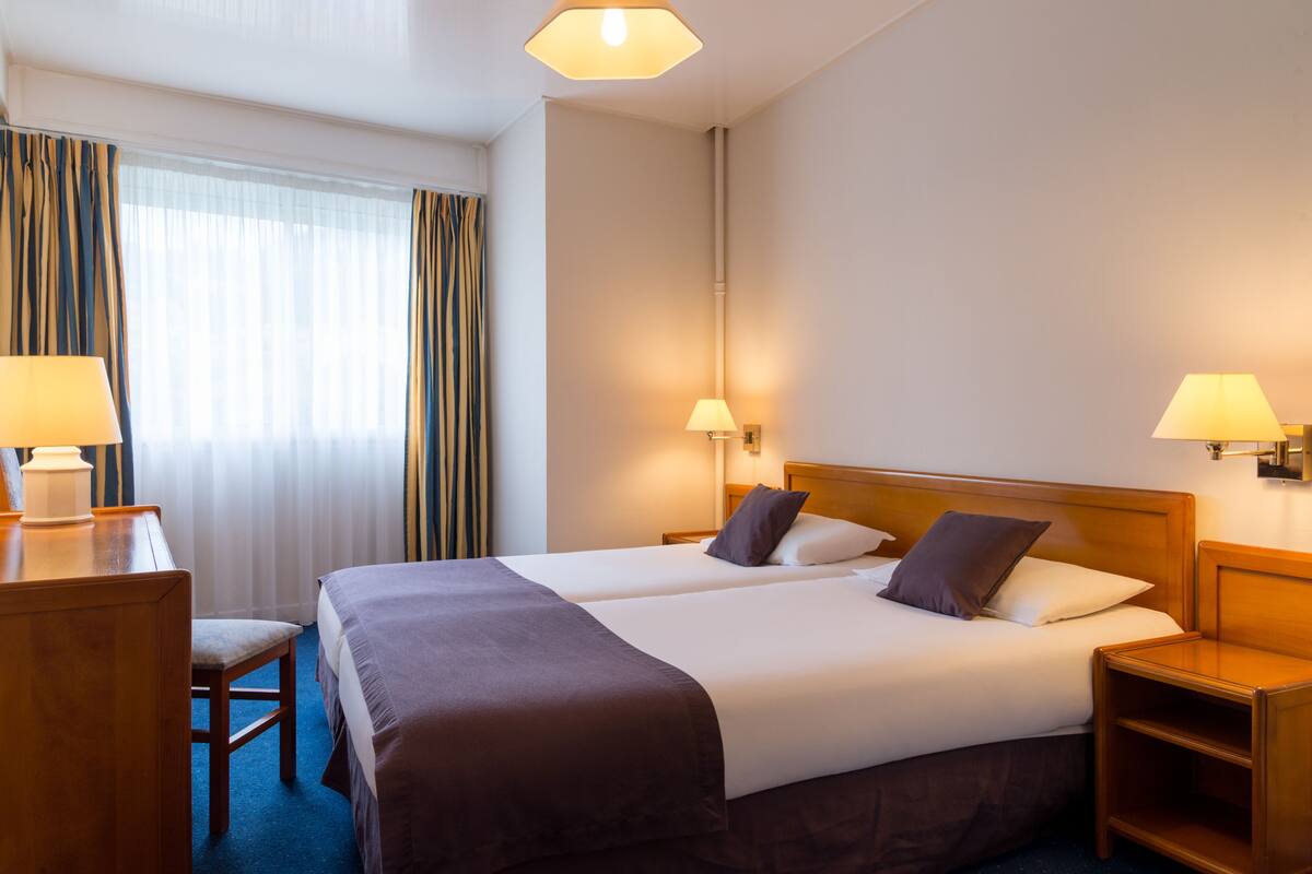 A hotel room with two beds, a desk, and a chair. There are lamps on either side of the beds and a window with curtains.