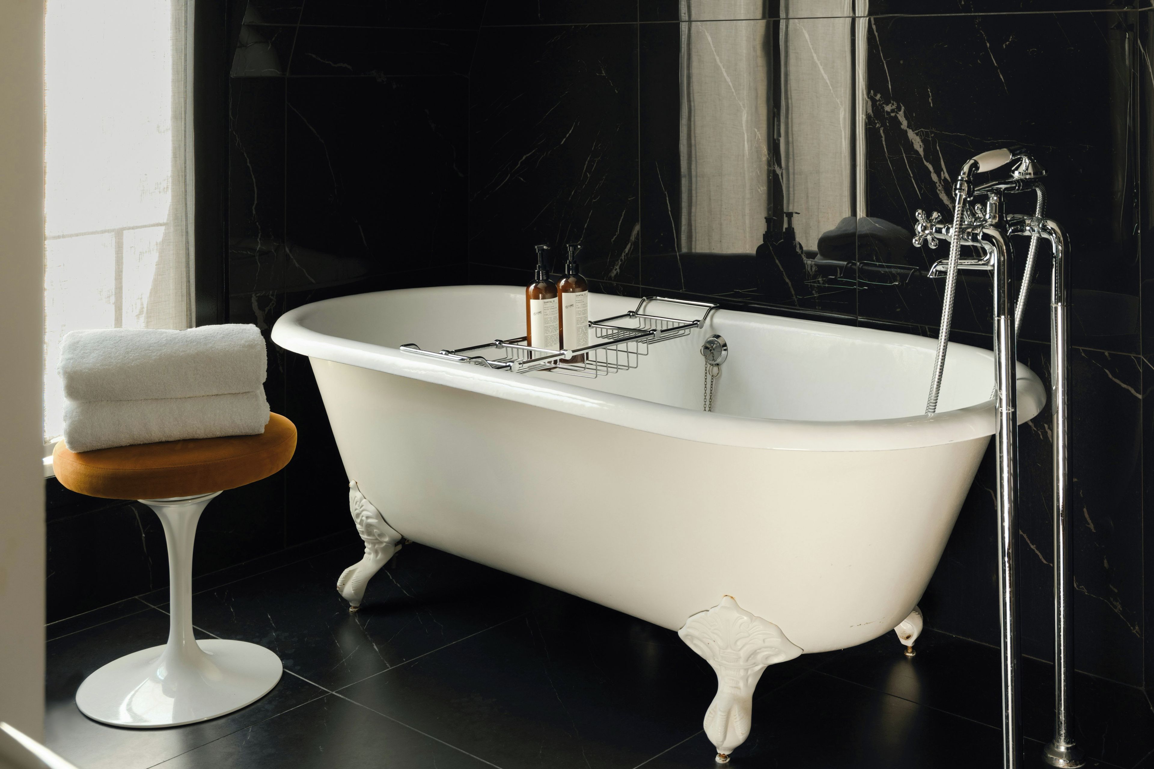 Eine freistehende Badewanne mit chromfarbenen Wasserhahn und Duschkopf, ein Handtuch auf einem Hocker und eine Wand mit Marmorfliesen.