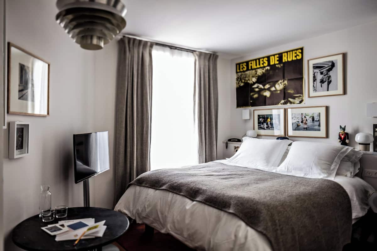 Una habitación de hotel con una cama, un televisor, una mesita con vasos y papeles, y un cartel en la pared.