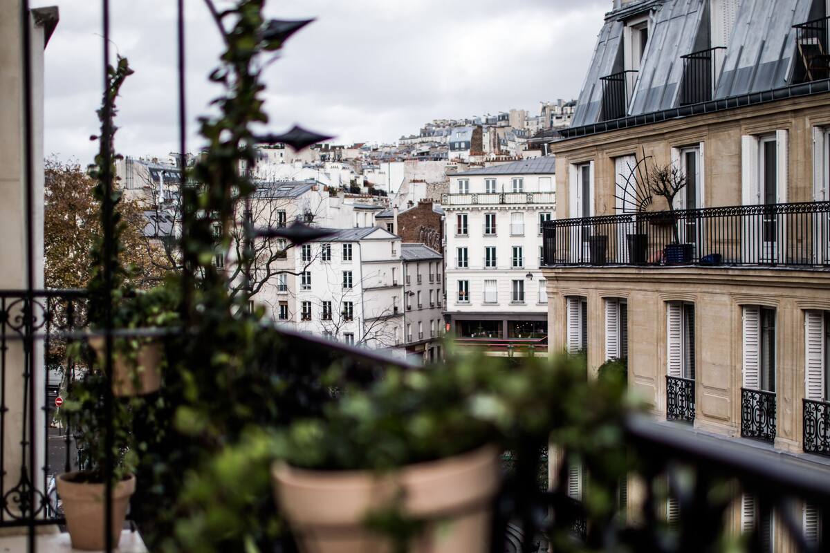 A balcony with potted plants, a metal chair, and a view of city buildings.