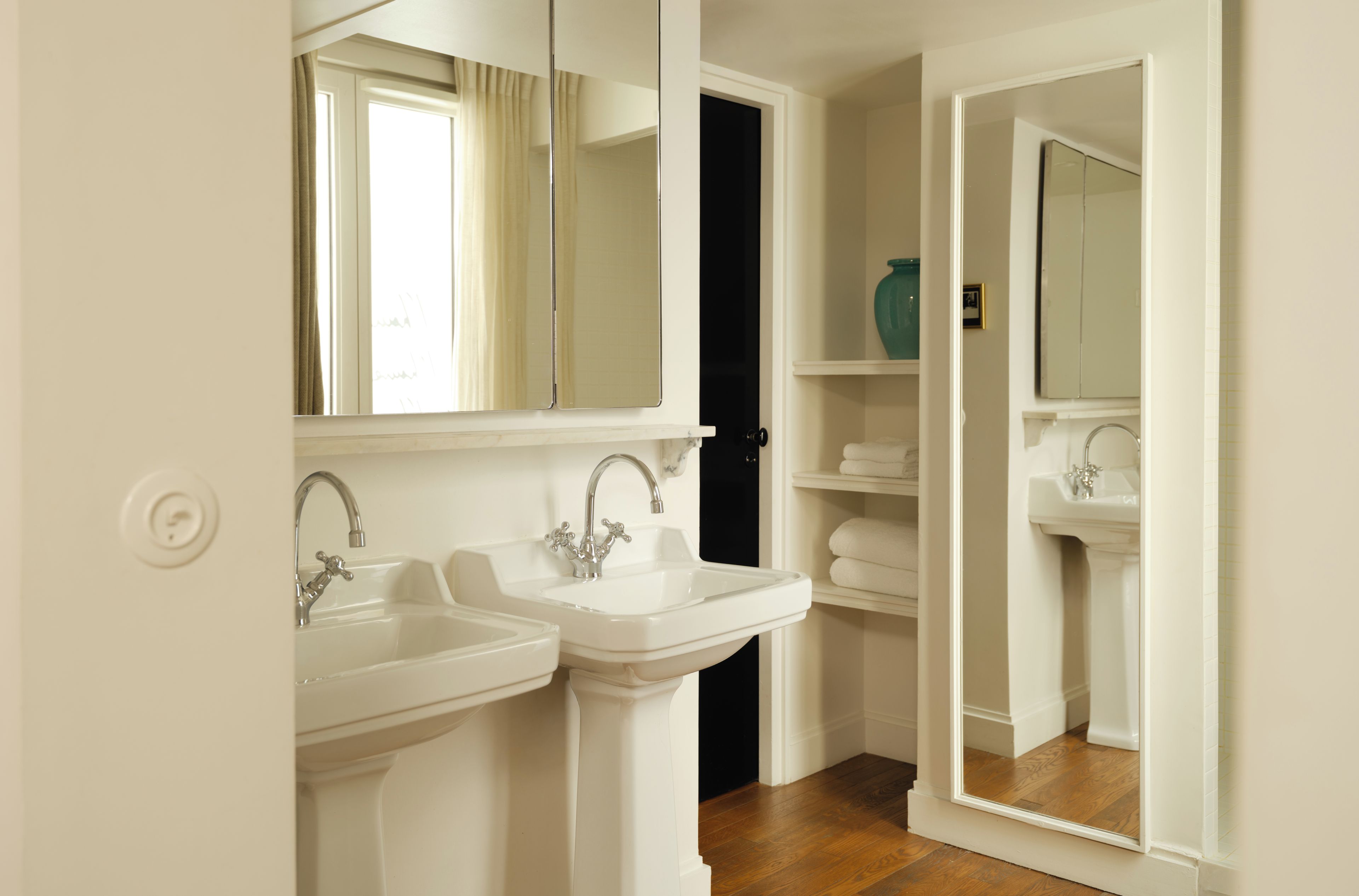 Un baño con dos lavamanos, un espejo y una puerta que da a otra habitación.