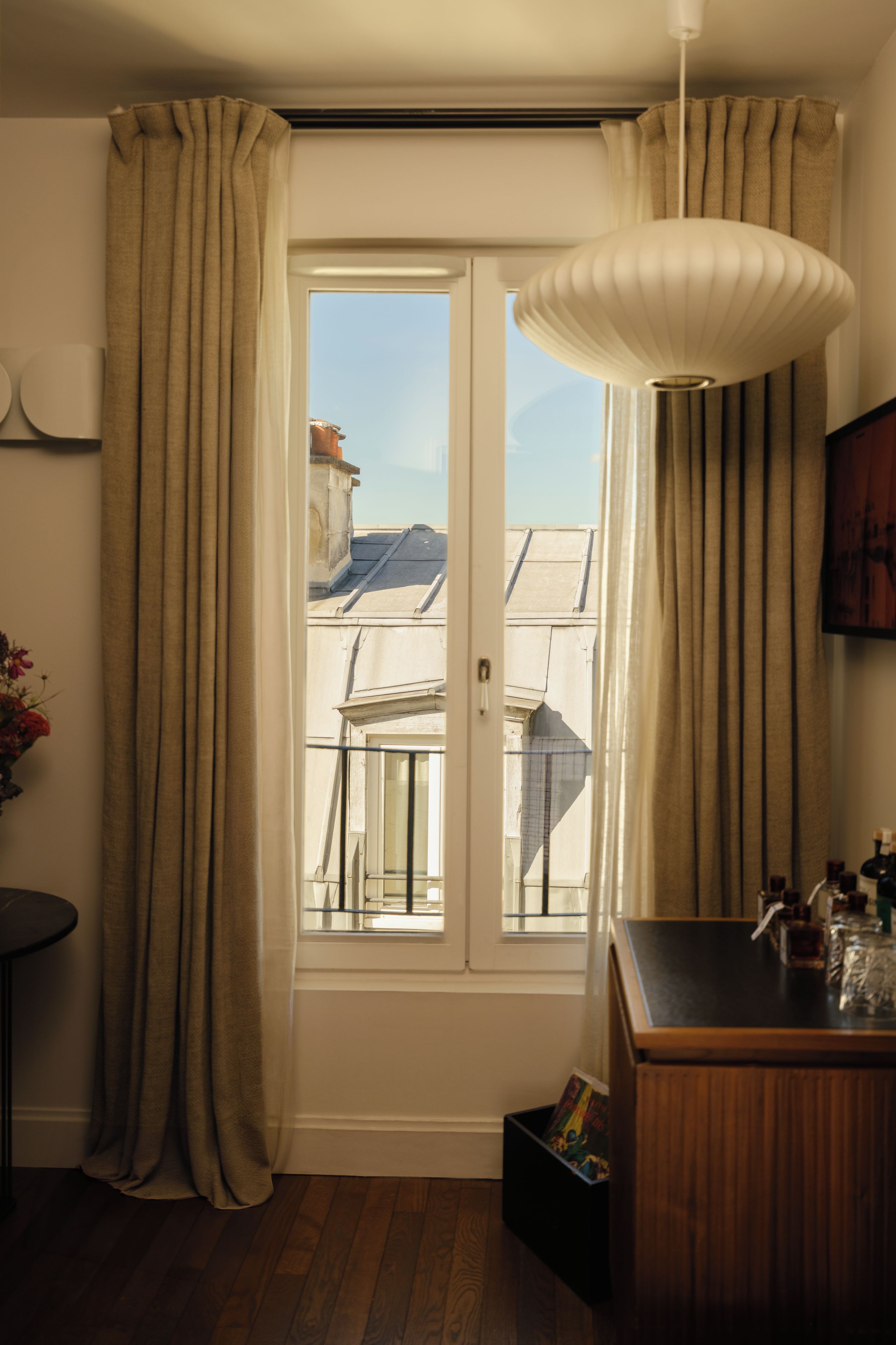 Een kamer met een deur die leidt naar een balkon, een kleine tafel met een bloevas, een houten kast met flessen en een hanglamp.