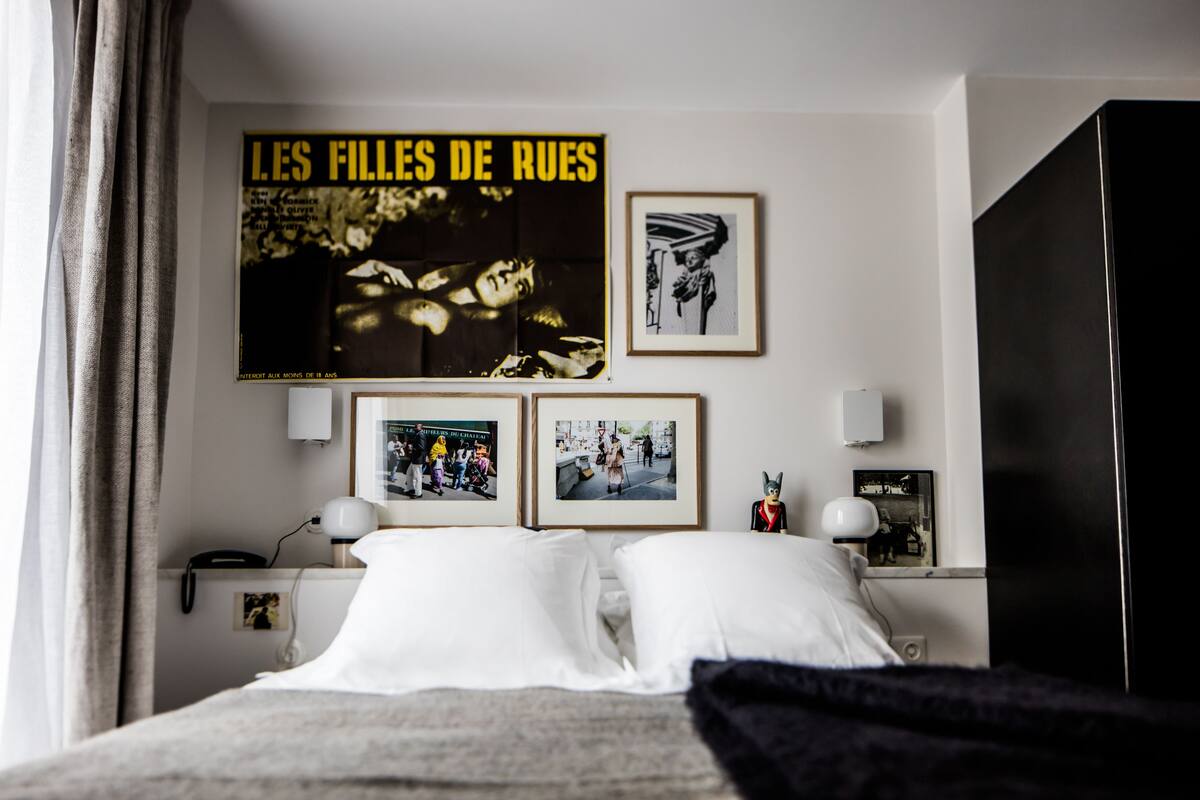 A hotel room with a bed, a poster on the wall, framed photos, and a nightstand with a lamp.