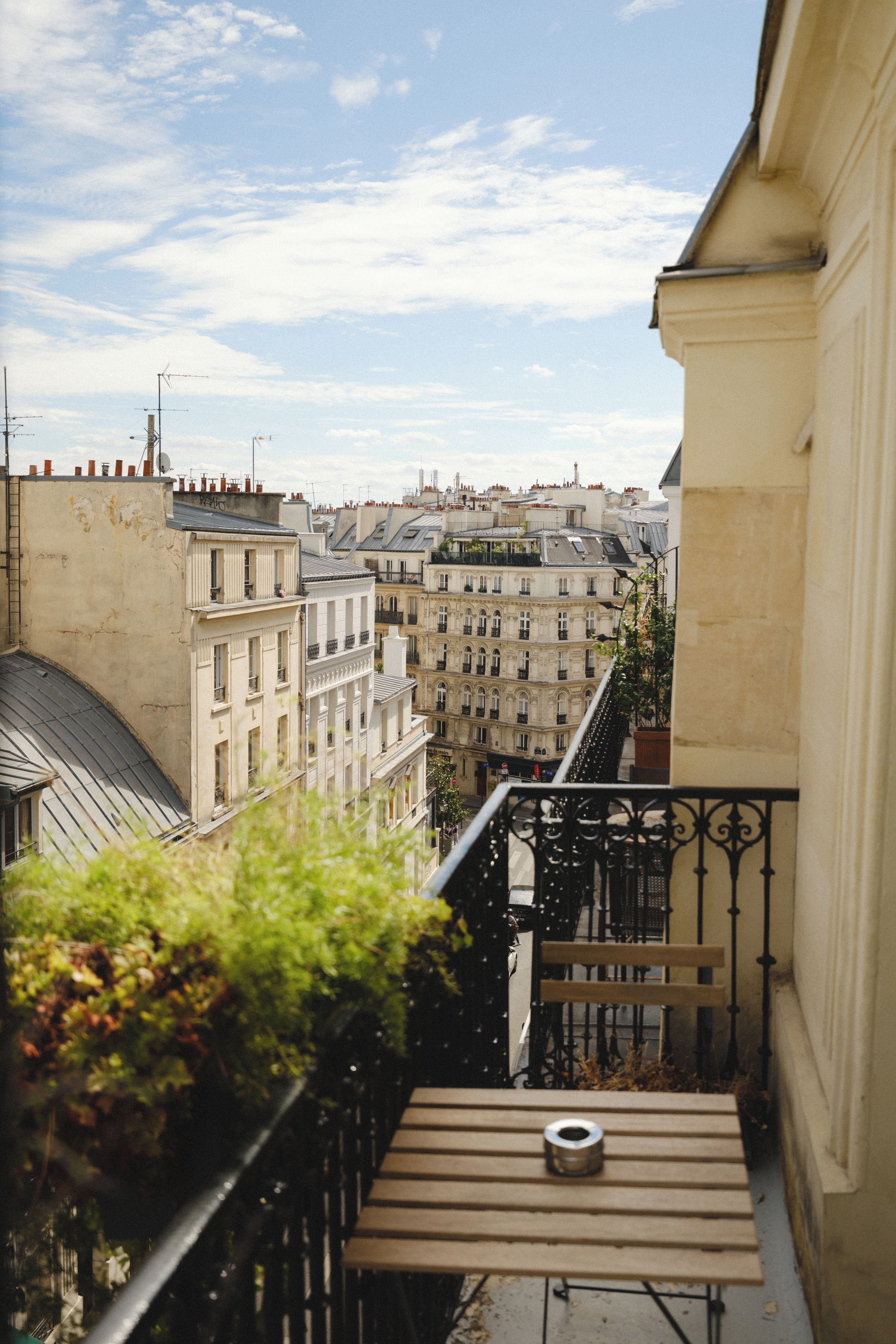 Un balcon avec une table et un pot de fleurs, donnant sur un paysage urbain avec des bâtiments historiques.