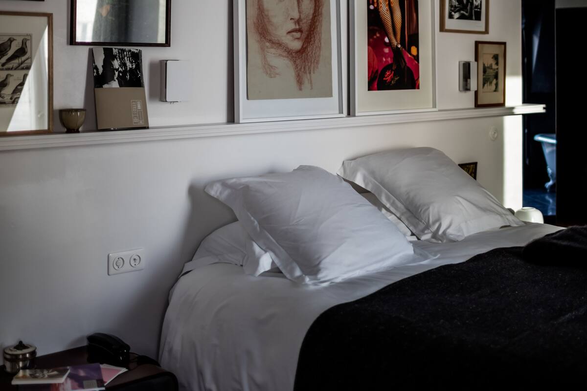 A bedroom with a bed, white pillows, and a black blanket. There are framed artworks on the wall and a nightstand with a lamp and a book.