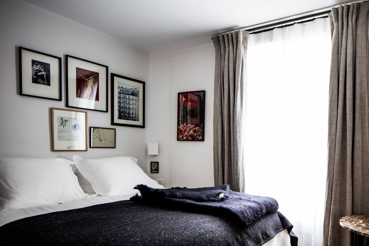 A hotel room with a neatly made bed, a window with curtains, and several framed pictures on the wall.