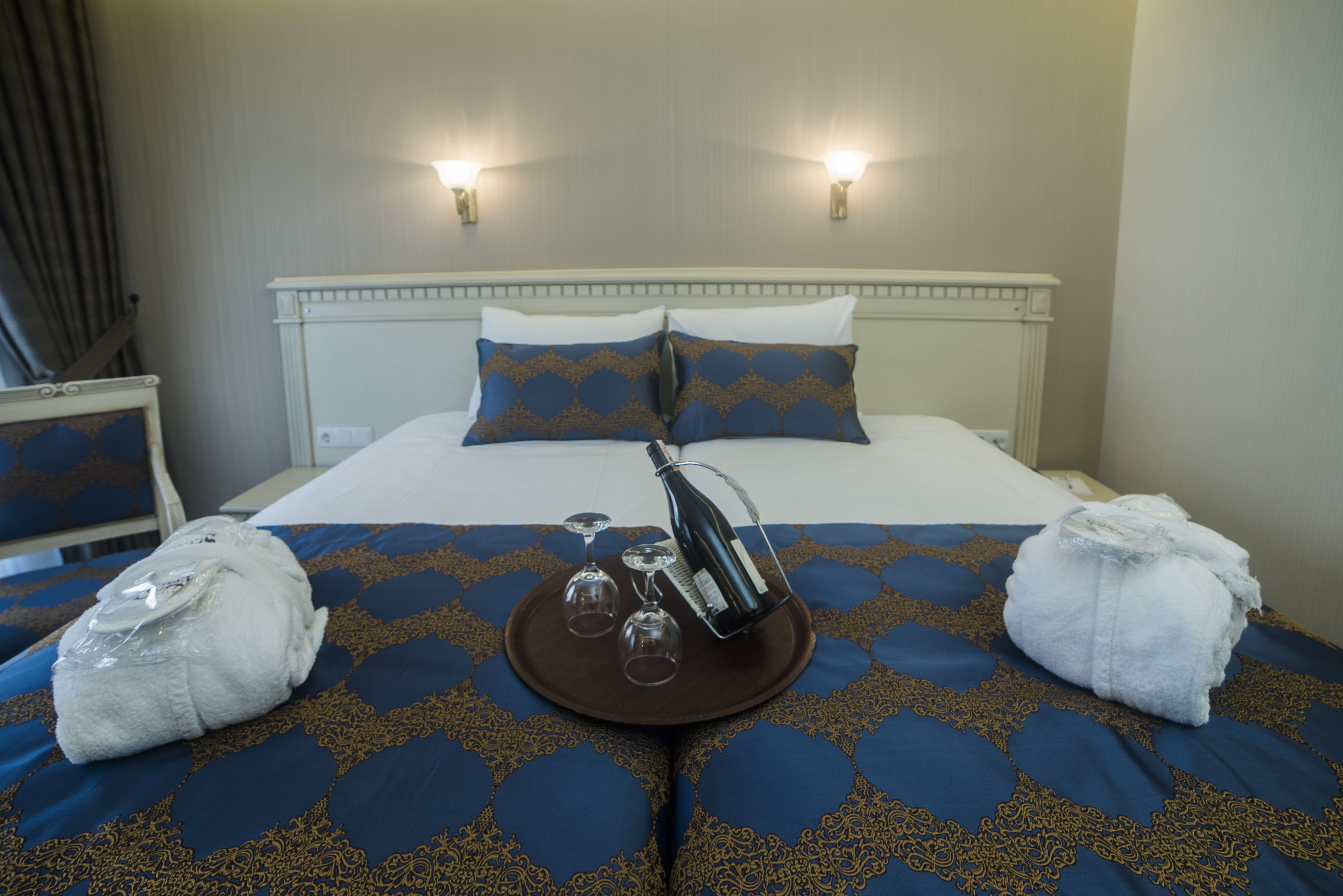 A hotel room with a bed, two wall-mounted lights, a tray with a bottle and glasses, and two towels on the bed.