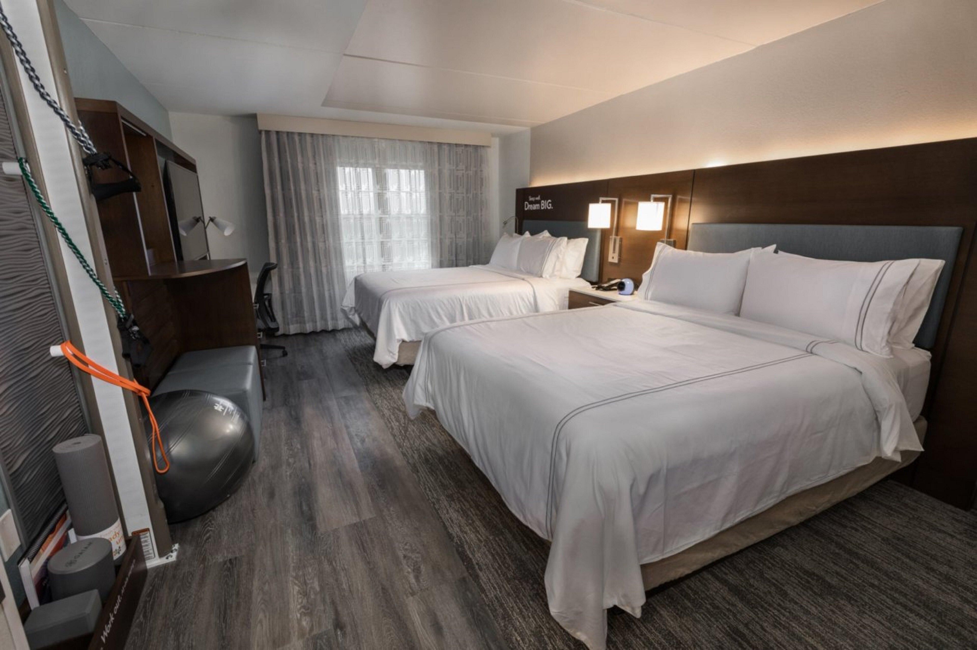 Habitación de hotel con dos camas, un escritorio, una silla, una cinta de correr y un espejo grande.