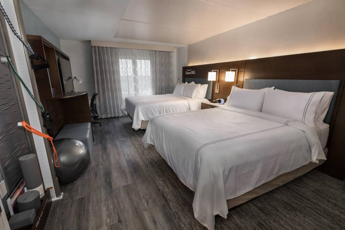 Habitación de hotel con dos camas, un escritorio, una silla, una cinta de correr y un espejo grande.