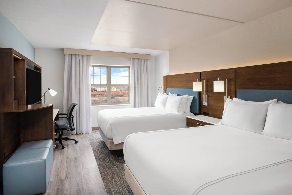 A hotel room with two beds, a desk with a chair, a TV, and a large window with curtains.