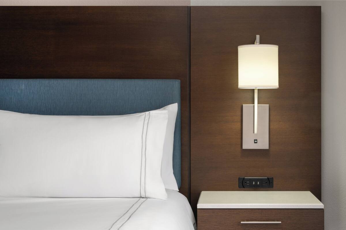 A hotel room with a bed, a nightstand with a lamp, and a wall-mounted power outlet.