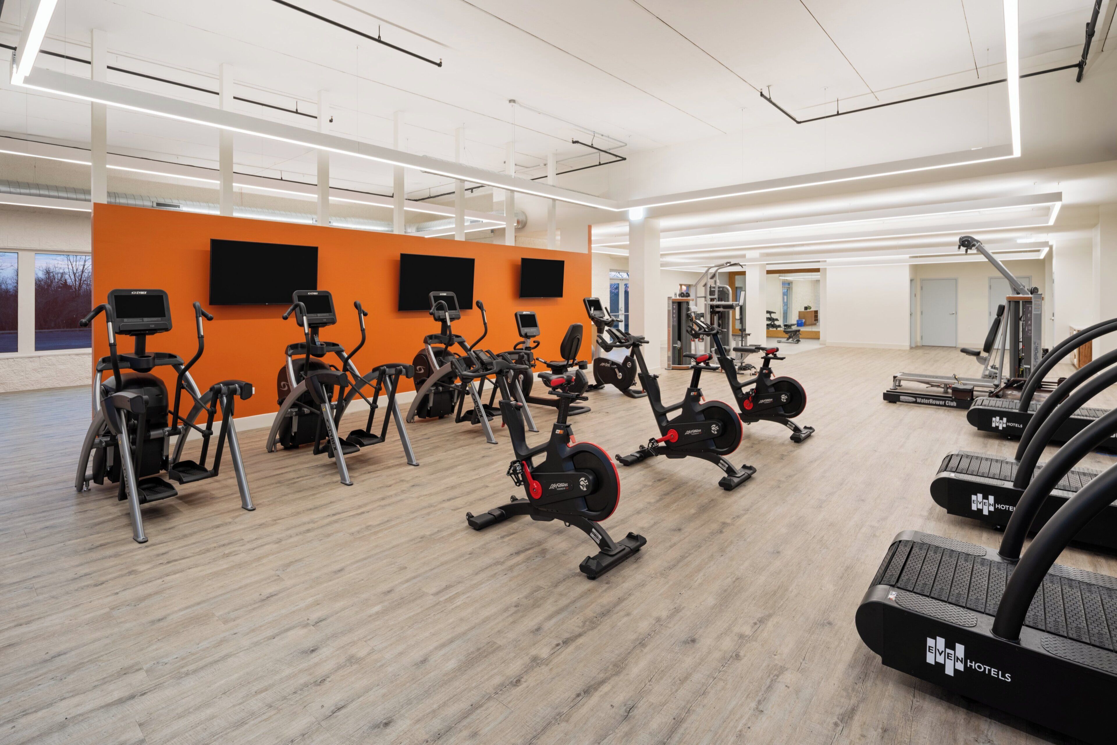 Et fitnessrum med forskellig træningsudstyr, herunder løbebånd og spinningcykler, i et rummeligt lokale med en orange væg og store vinduer.
