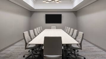 A conference room with a long table, white chairs, a flat-screen TV, and a skylight ceiling.