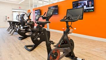 Une rangée de vélos d’appartement dans une salle de sport aux murs orange et au plancher en bois.