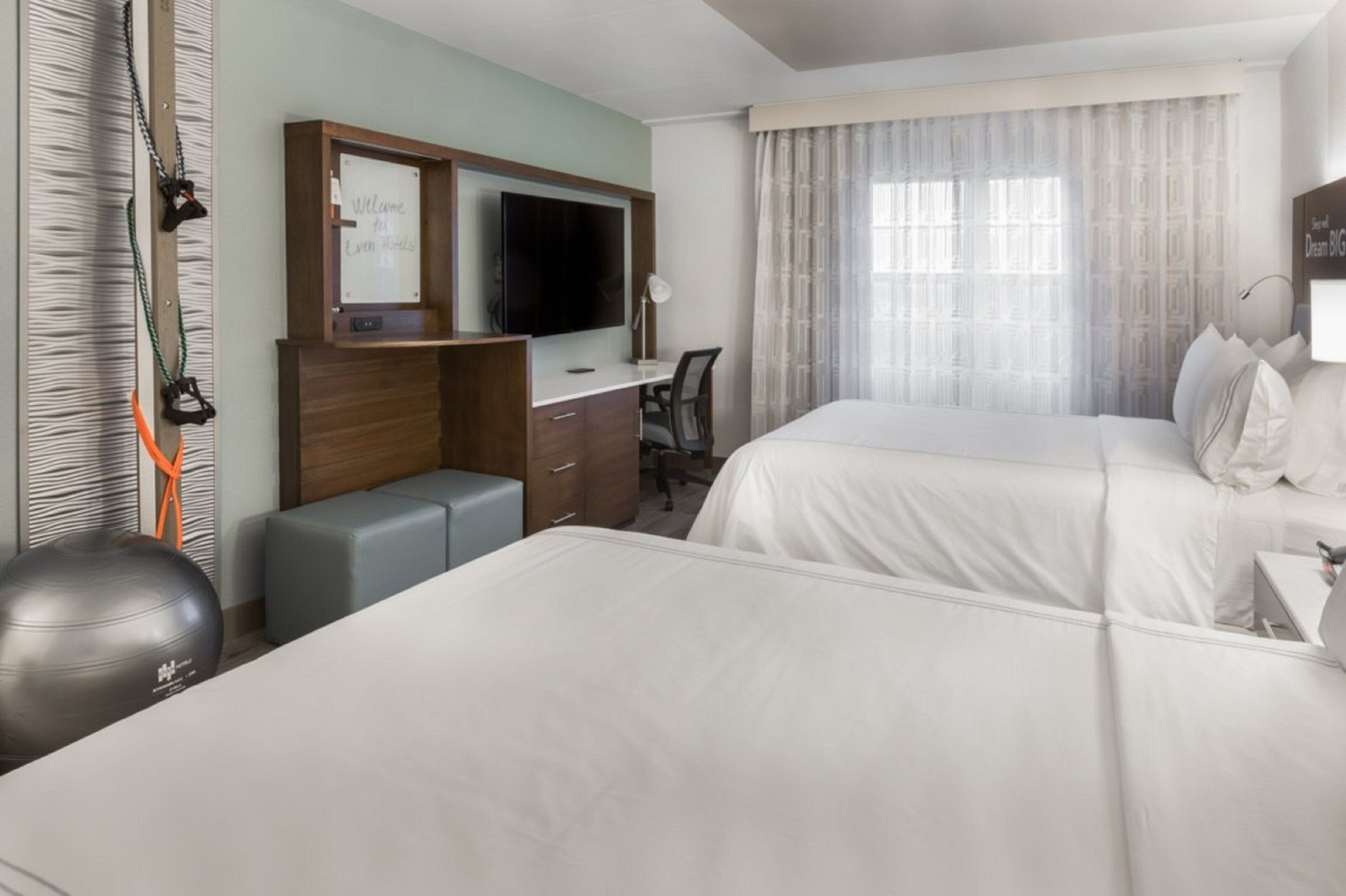 A hotel room with two beds, a desk, a television, exercise equipment, and a large window with curtains.