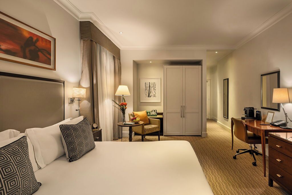 A hotel room with a large bed, a desk, a chair, a small table with a lamp, and a wardrobe.