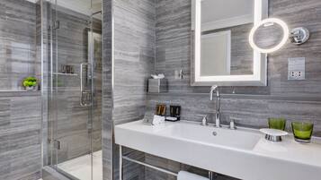 Un baño moderno con ducha envuelta en vidrio, un espejo grande con luz integrada y un lavabo blanco con vasos de vidrio verde.