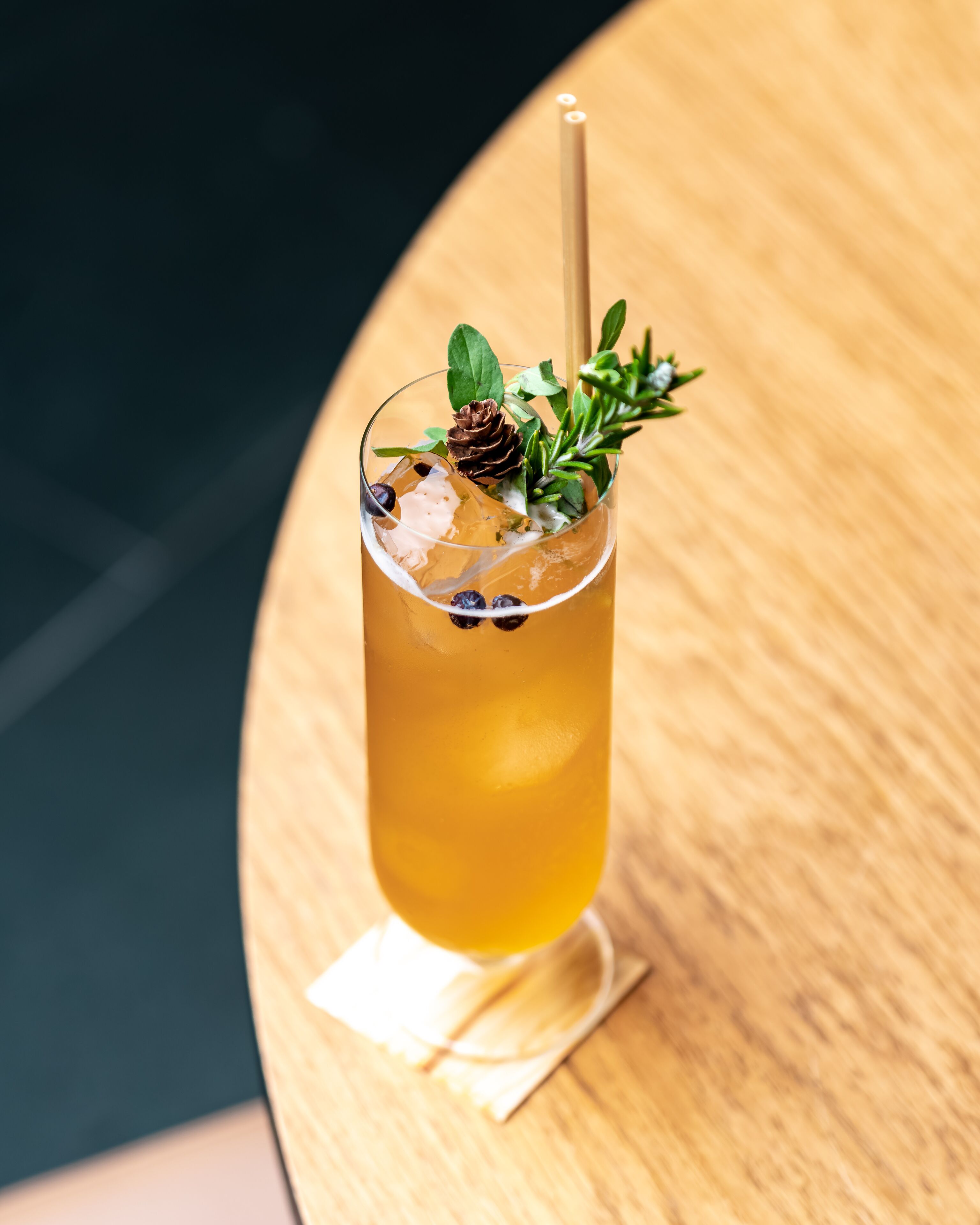 A tall glass with a straw, garnished with pine cones and herbs, on a wooden table.