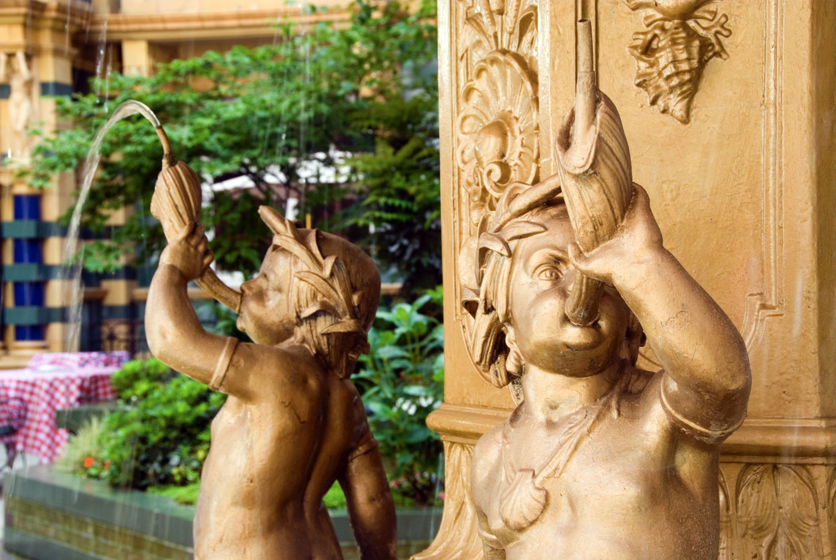 Eine goldene Statue einer Gestalt mit erhobener Arm und flatterndem Gewand, vor dem Hintergrund von Grünpflanzen und architektonischen Details.