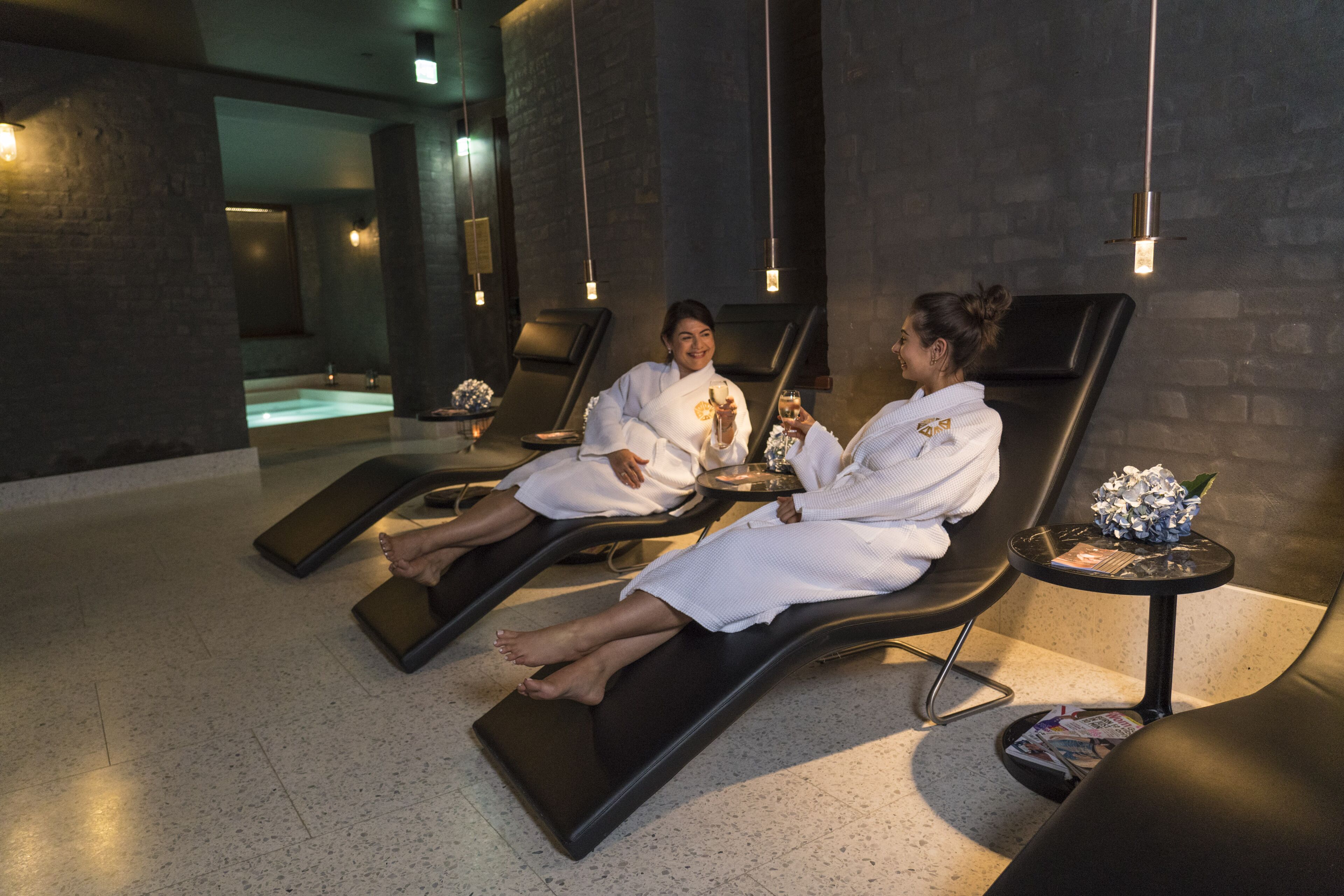 A spa room with two reclining chairs, a small table with a magazine, and a wall-mounted light fixture.
