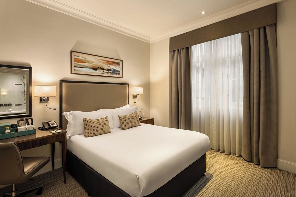 A hotel room with a large bed, a desk with a telephone, a chair, and a window with curtains.