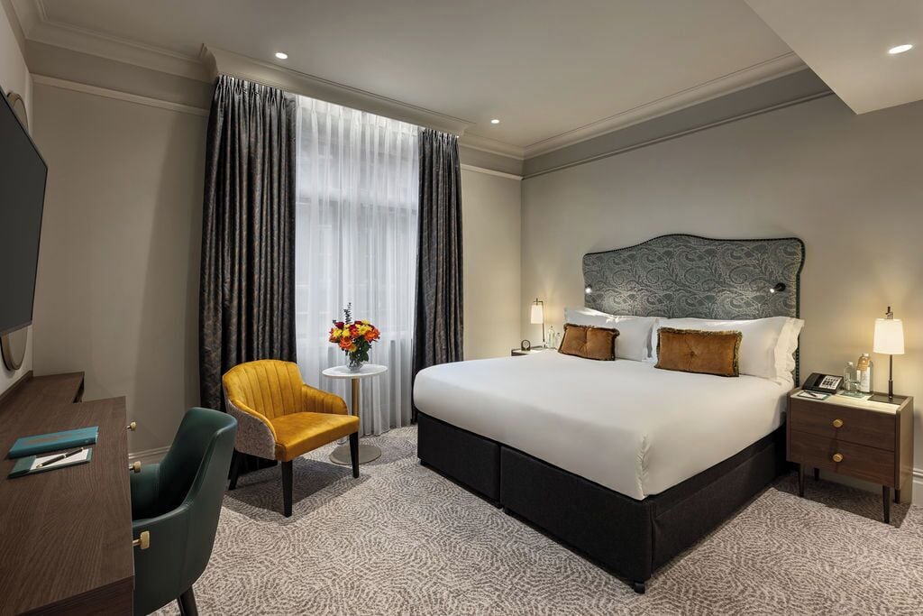 A hotel room with a large bed, a desk with a chair, a small table with a vase of flowers, and a nightstand with a lamp.
