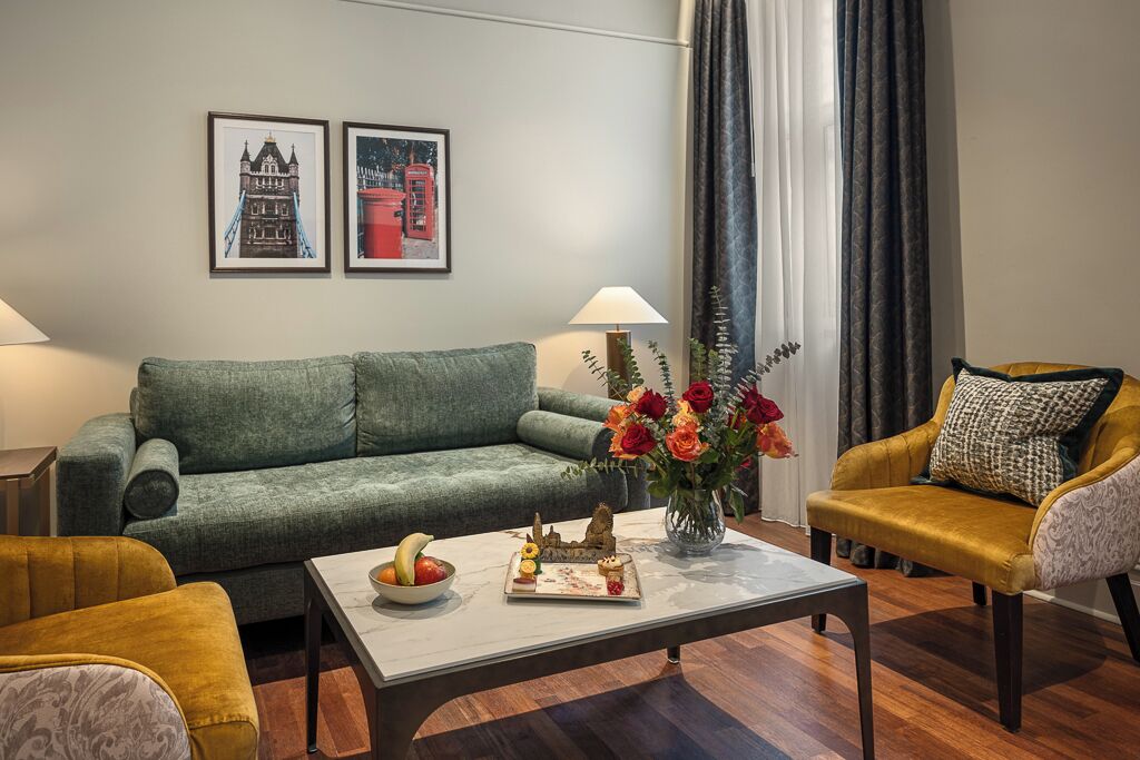 A living room with a green sofa, a yellow armchair, a white coffee table with fruit and a decorative item, and two framed pictures on the wall.