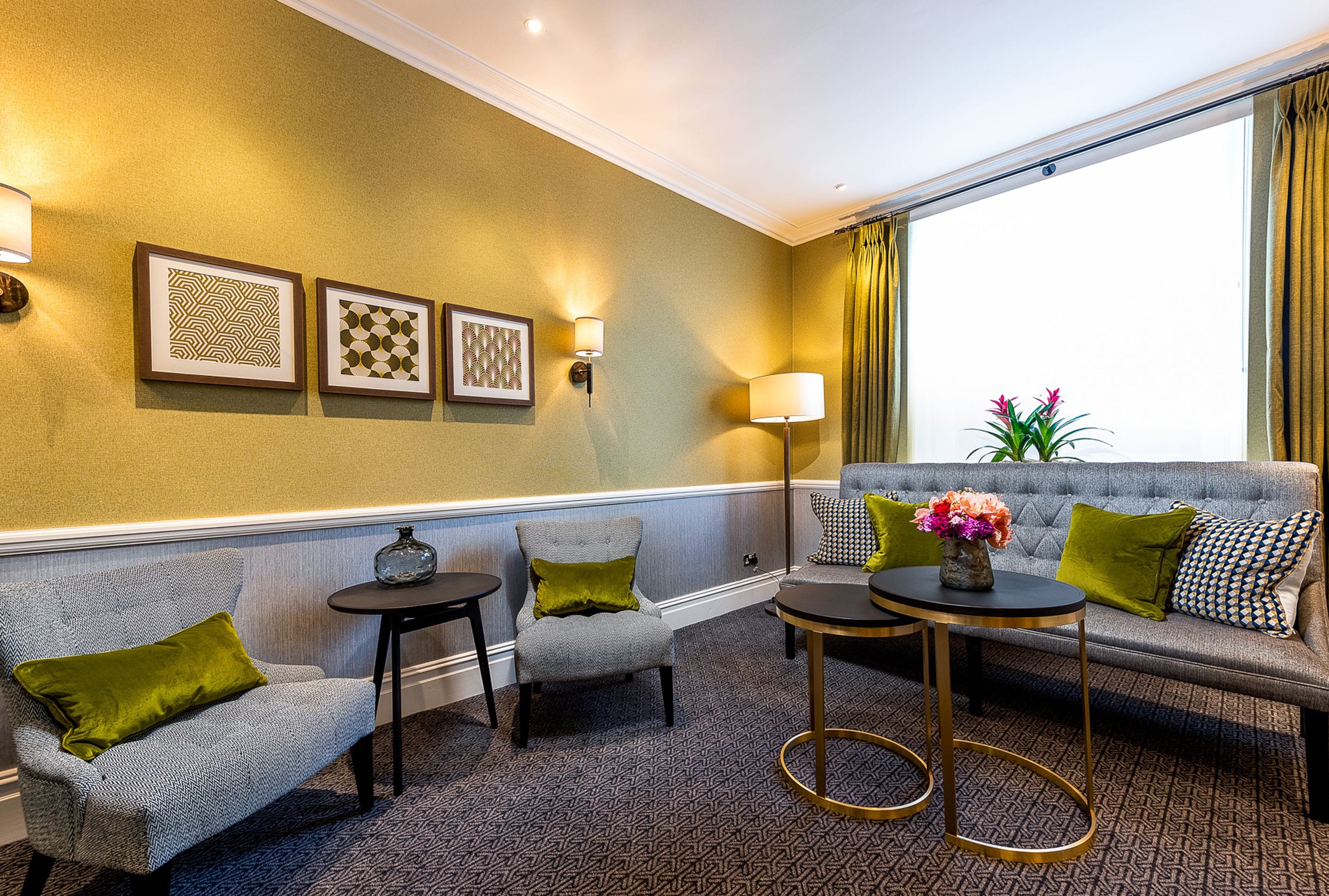 A hotel room with a grey sofa, two armchairs, a small round table, and a floor lamp. There are framed artworks on the wall and a window with curtains.