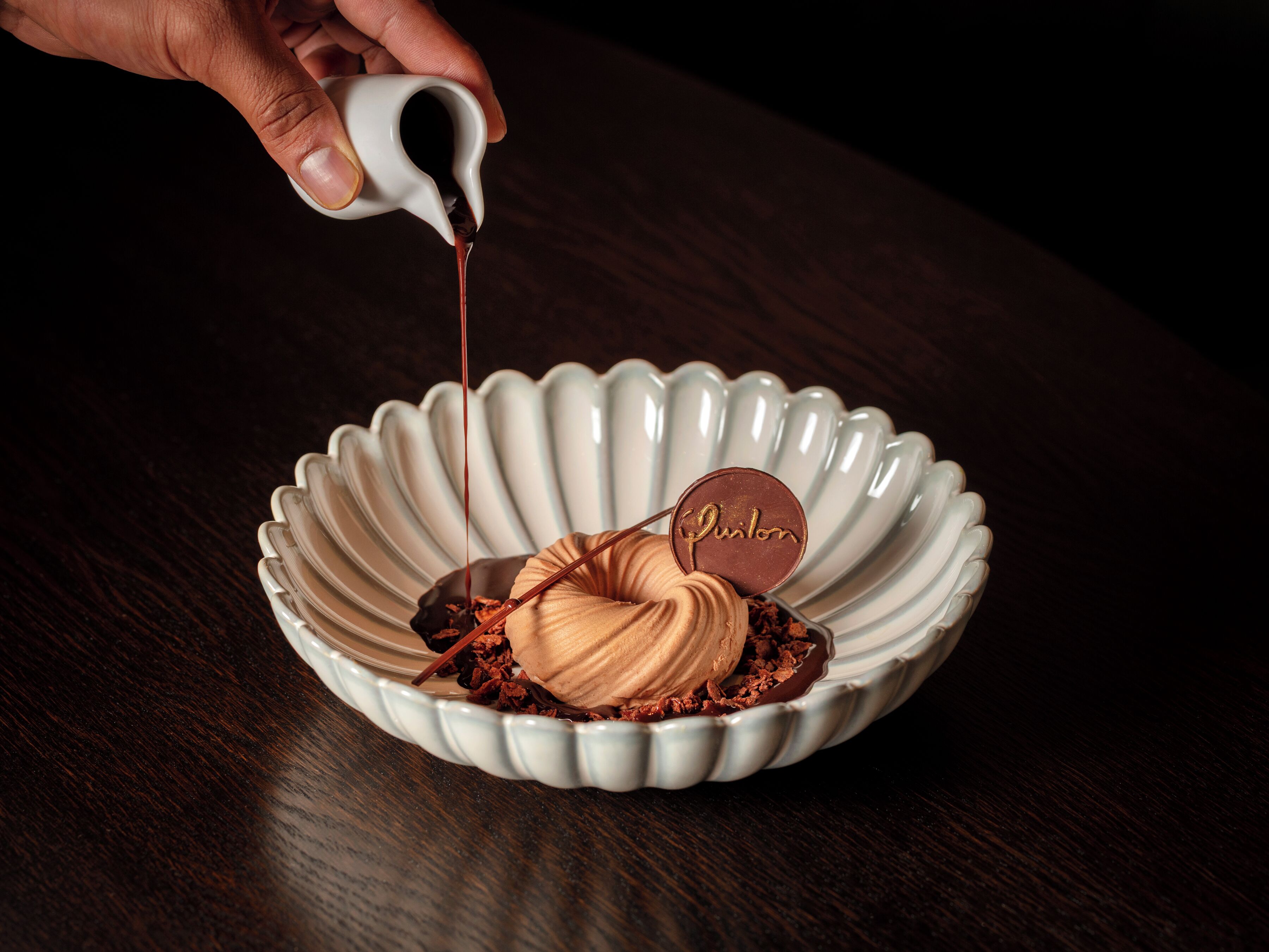 A hand pouring chocolate into a bowl with a chocolate dessert inside.