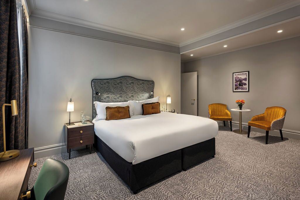A hotel room with a large bed, two bedside tables with lamps, a small round table with flowers, two armchairs, and a framed picture on the wall.