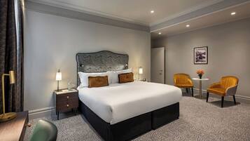 A hotel room with a large bed, two bedside tables with lamps, a small round table with flowers, two armchairs, and a framed picture on the wall.