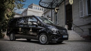 Un fourgon Mercedes-Benz Sprinter noir était garé devant un hôtel au style architectural classique, avec une entrée imposante et une enseigne indiquant qu’il s’agissait d’un hôtel.