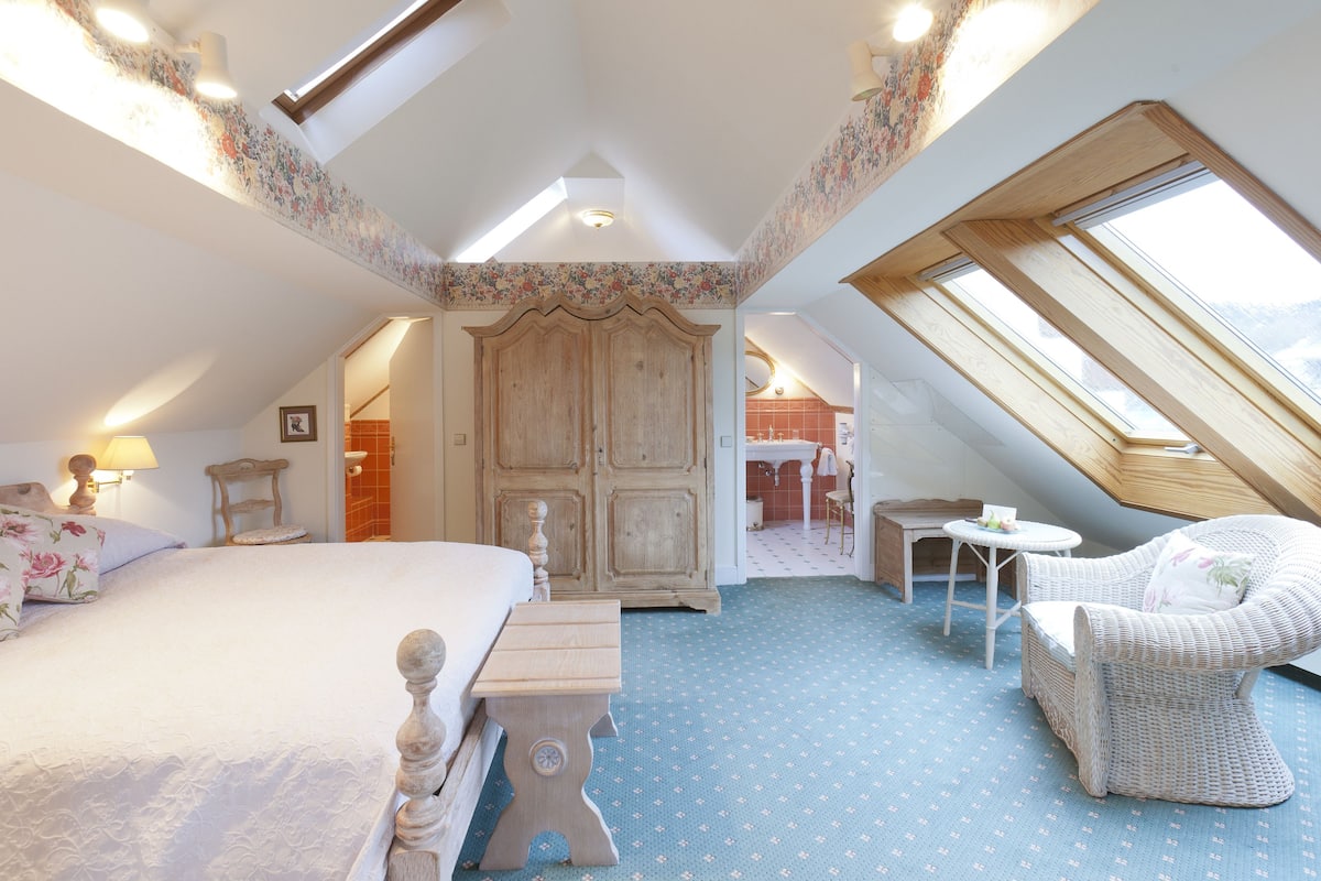A bedroom with a large bed, a wooden wardrobe, a wicker chair, a small table, and a skylight.