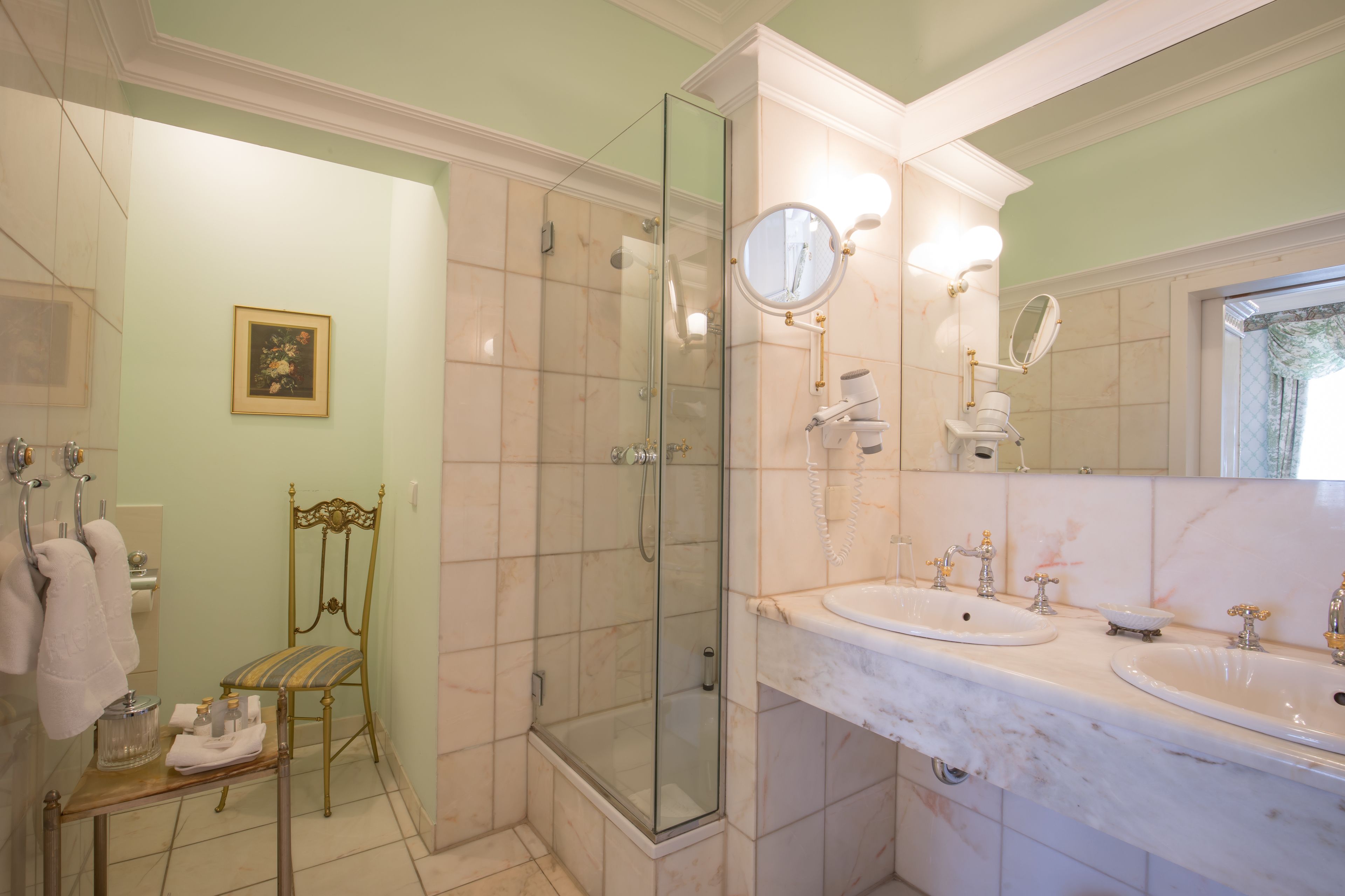 Un baño con ducha envuelta en vidrio, dos lavamanos, un espejo sobre el mueble y una silla con cojín.