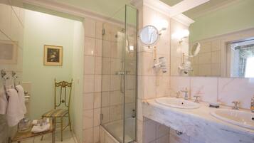 Un baño con ducha envuelta en vidrio, dos lavamanos, un espejo sobre el mueble y una silla con cojín.