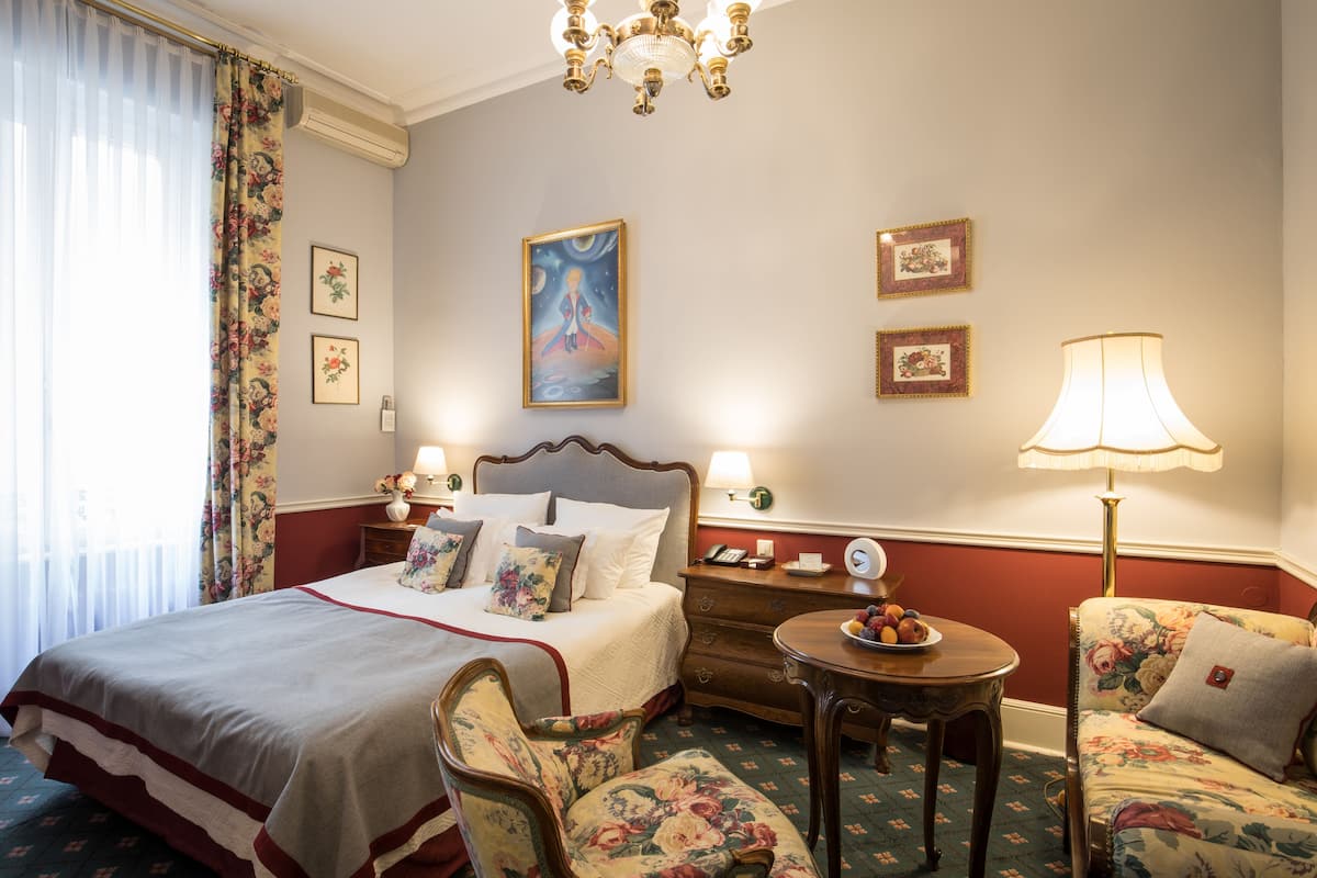 Une chambre d’hôtel avec un grand lit, des fauteuils à motifs floraux, une petite table et un lustre.
