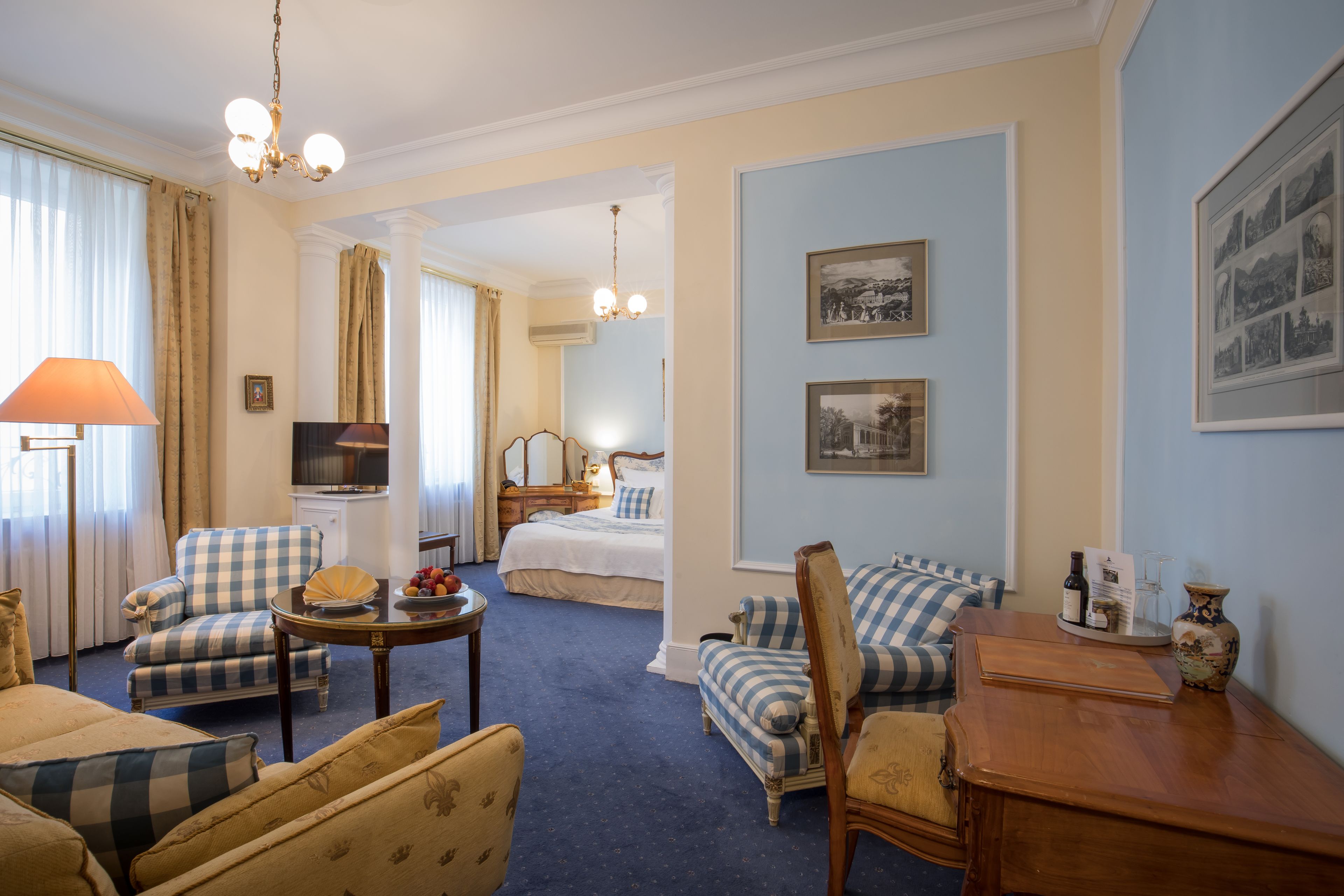 A hotel room with a checkered armchair, a sofa, a dining table, a lamp, a bed, and framed pictures on the wall.