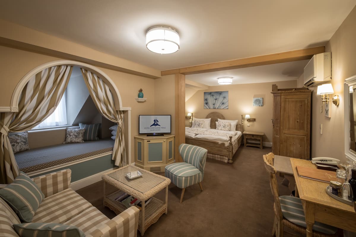 Une chambre d’hôtel comprenant un lit, une télévision, une table à manger, une chaise, un canapé et une fenêtre avec des rideaux.