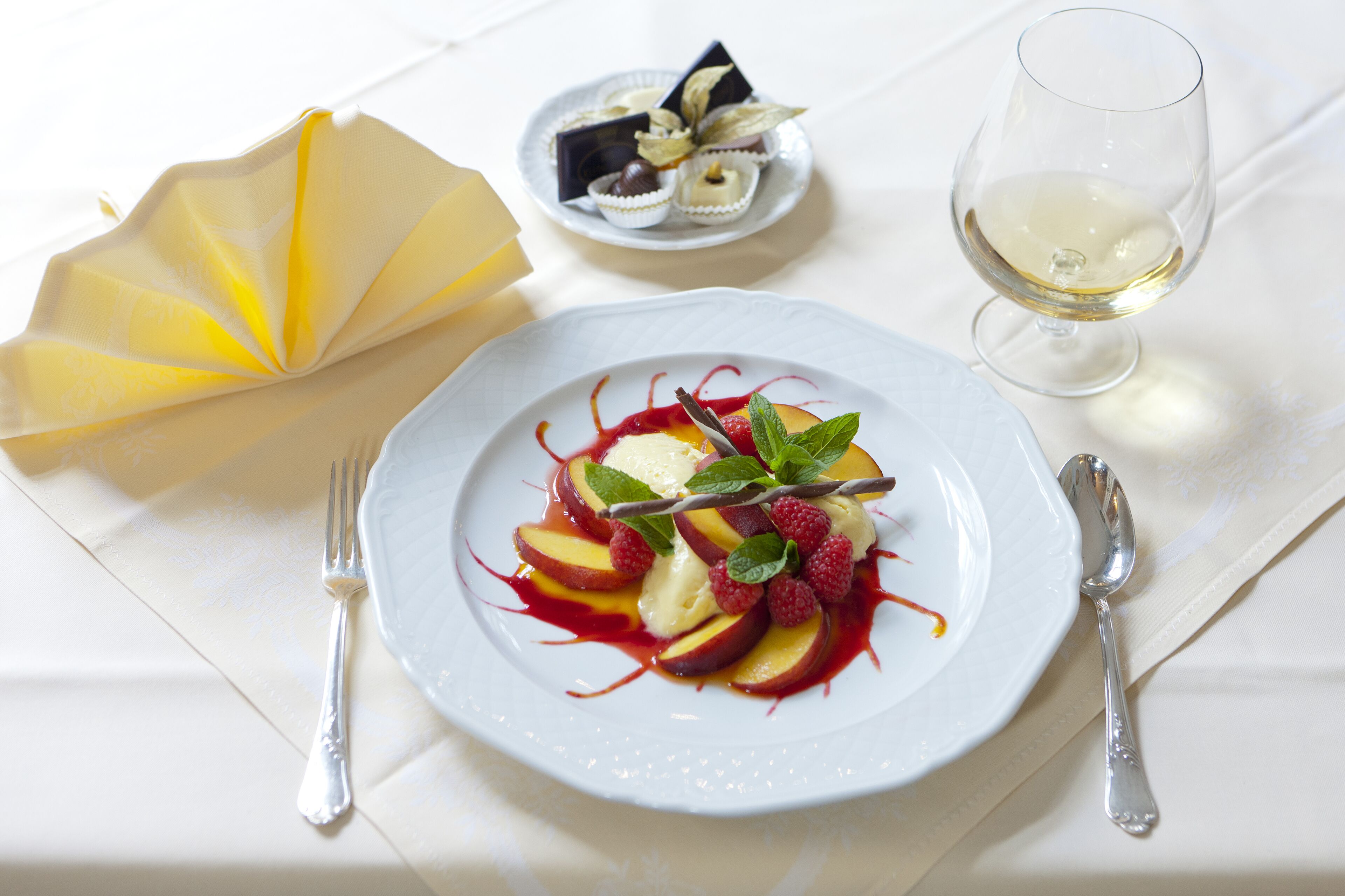Un plato de postre con fruta y menta, un pequeño plato de bombones, una copa de vino blanco y cubiertos sobre un mantel blanco.