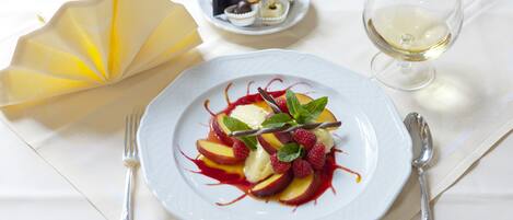 Un plato de postre con fruta y menta, un pequeño plato de bombones, una copa de vino blanco y cubiertos sobre un mantel blanco.